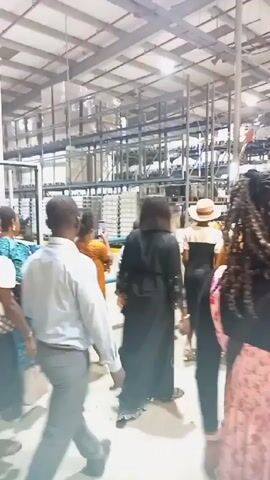 A group of people walk through a large warehouse or factory setting. High shelves filled with goods line the walls. The group passes a large, white electrical panel with vents. They are walking behind a blue railing. Some members of the group are talking and gesturing. The environment appears brightly lit and orderly.