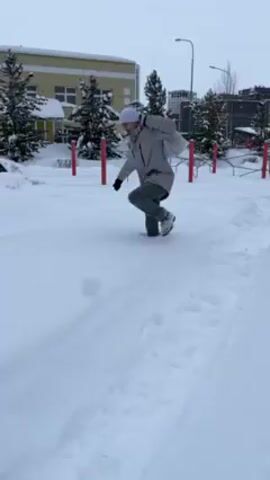 The video shows a person attempting to walk through deep snow in an outdoor setting. The person is wearing a light-colored winter coat, dark gloves, and a white beanie. They appear to be struggling as they take each step, sinking deeply into the snow. The setting appears to be a park or playground covered in snow, with trees, a building, and playground equipment visible in the background. The ground is heavily covered in untracked snow, except for a path someone previously walked.