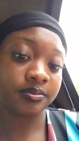 A close-up selfie-style video captures a young Black woman with dark skin, wearing dark lipstick and a black headwrap. She has prominent eyelashes. Initially, her expression is neutral. Approximately two seconds into the video, she smiles broadly, revealing her teeth. A light-colored substance is visible on the inner part of her lower lip. She is wearing a multi-colored top with stripes of blue and black, and a block of maroon. The background is a blurry, overexposed light area. An audible wind-like sound persists throughout the video.