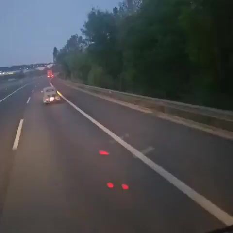 A light green, vintage Renault 4CV car is traveling on a highway. The road is dark, with a white line separating lanes. The right side of the road has a metal guardrail and trees in the background. The car's rear lights are illuminated.  The video is shot from a vehicle moving behind and to the left of the Renault.