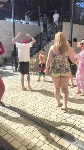 In the video, a group of people are gathered outdoors on a paved area. A person dressed in a Deadpool costume is dancing and interacting with others. A man in a white shirt and red cap is also dancing. A woman in a floral dress is standing near the center, and another woman in a black top is clapping. A young boy is standing between the dancing man and the woman in the floral dress. In the background, there is a staircase, some seating, and trees. Later in the video, the scene shifts to a different angle showing a group of children and adults near a wooden deck. A young girl wearing a blue inflatable float is looking to the side. The weather appears to be sunny.