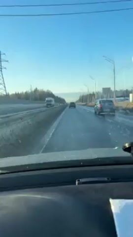 The video, captured from inside a vehicle, shows a view of a highway under a bright, clear, blue sky. The road is mostly clear, though some patches of wetness are visible.  A truck is present in the left lane, and a car is driving in the right lane.  Other vehicles are scattered further down the road. The scenery on either side of the highway includes trees and utility poles. The dashboard of the car from which the video is recorded is visible in the foreground.