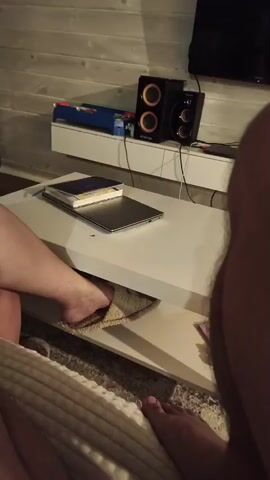 The video shows a close-up view of a white coffee table with a laptop and books on it. A person's bare foot, wearing a slipper, is visible on the lower shelf of the table. In the background, there are speakers and a television mounted on a wooden wall. The scene appears to be in a living room or a similar domestic setting.