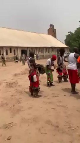 In a medium shot, a group of six men perform a cultural dance in a sandy courtyard, against the backdrop of a church with a tall, square tower. The men are all wearing white tank tops, with the lower half of their bodies adorned in vibrantly colored traditional clothing. One of the men carries a large, bushy object made of natural materials. The dancers move in a circle, their faces visible and expressing joy. The church in the background is light-colored, with a simple design. In the background, other people and parked cars can be seen. The environment appears to be rural or suburban, with trees framing the scene. The sky overhead is overcast.