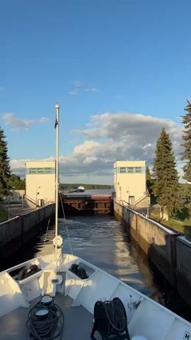 The video shows a view from a boat approaching a lock system. The boat is moving forward into the lock. Two white buildings with blue trim stand on either side of the lock entrance. A large, brown metal gate is visible ahead, partially open. Beyond the gate, a calm body of water is seen, with a blue and white ship sailing in the distance. A red car is driving across a red bridge that spans the lock system in front of the open gate. The sky is blue with fluffy white clouds, and green trees line the horizon. The overall impression is a functional waterway system in a natural setting under a clear sky.