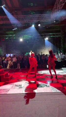 Two women are dancing on a checkered floor in a dimly lit venue. The woman on the left is wearing a bright pink dress with fringe details and black heels. The woman on the right is wearing a green sequined dress and black heels. Both women have their arms raised as they dance. In the background, a crowd of people are watching the performance. Stage lighting illuminates the dancers and the checkered floor.