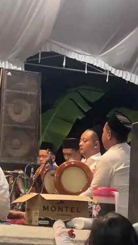 The video shows a group of men performing music. They are wearing traditional Islamic clothing, including white shirts and black caps. One man is playing a stringed instrument, possibly a violin, while others are playing percussion instruments, likely drums. There are large speakers visible in the background, suggesting a public performance or event. The scene appears to be set up outdoors, possibly under a tent, with draped white fabric overhead and green foliage in the background. A person wearing a hijab is partially visible in the foreground, looking towards the musicians. There are also some drinks and a box with the brand name "MONTEL" on it on the ground. The lighting is dim, indicating it might be evening or night.