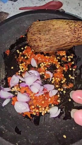 A close-up shot shows a person's hand holding a halved lime over a dark gray mortar containing chopped red chilies and sliced red onions. A wooden pestle rests inside the mortar, and the chilies and onions appear to be partially mashed. The hand is gently squeezing the lime, allowing juice to drip into the mortar. The background is blurred, but hints of a countertop and a red spoon are visible.