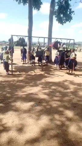 A group of children, mostly girls in school uniforms, are playing on a swing set in a sandy, outdoor area. The swing set is made of metal poles and bars. Two children are actively swinging, with one appearing to be mid-air. Other children are standing around the swing set, some watching, some waiting for their turn, and a few are climbing on the frame. The area is dusty with some patches of dry leaves on the ground. Two tall trees with large canopies are visible behind the swing set, providing shade. The sky is blue with scattered white clouds. In the background, there are some buildings and a distant hillside. The overall impression is a schoolyard during playtime on a sunny day.