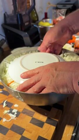 A person's hands are shown pressing a white plate down onto shredded cabbage in a large metal bowl. The bowl is on a wooden cutting board with a checkered pattern. Scattered pieces of cabbage are on the cutting board. In the background, a blender and other kitchen items are visible. The person is tamping down the cabbage.
