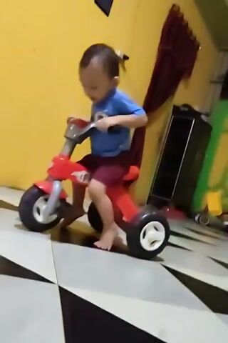 A blurry, low-light video shows a young child in a blue shirt and red shorts riding a red tricycle. The child's face is indistinct due to motion blur. The tricycle has three white-rimmed wheels. The child is seated on the tricycle, with their hands on the handlebars. The background consists of a yellow wall and a blue wall, with some indistinct objects leaning against the blue wall. The floor has a checkered pattern of light and dark squares. The video appears to be filmed indoors.