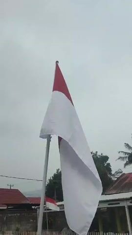 A red and white flag waves from a flagpole against a cloudy sky. The flag is mostly white with a red top portion. In the background, there are some buildings with red roofs and a wooden fence. A smaller red and white flag is visible in the distance. The scene appears to be outdoors in a somewhat rural or suburban setting.