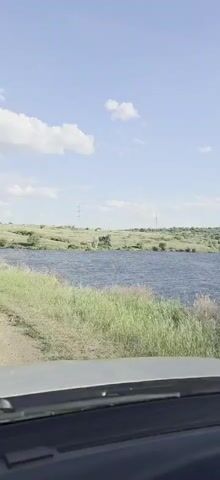 A car is driving next to a large body of water, possibly a lake or reservoir. The water is choppy, suggesting wind or waves. On the far side of the water, there is a gentle slope covered in green grass and trees. In the distance, there are several tall, thin structures that appear to be radio towers or power poles. The sky is blue with scattered white clouds, indicating it is daytime and likely fair weather. The camera is positioned inside the car, looking out through the windshield. The bottom of the windshield and dashboard are visible in the foreground. The video appears to be a short, continuous shot, possibly a snapshot from a longer journey.
