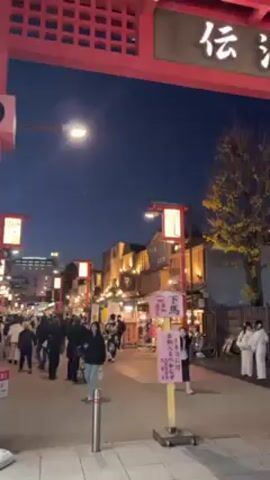 The video shows a night view of a crowded street in Japan. People are walking under a large red gate with Japanese characters on it, which is lit up. The sky is a deep blue. There are streetlights and lanterns illuminating the area. On the sides of the street, there are stalls and shops. Some people are wearing traditional Japanese clothing. The overall atmosphere is lively and festive.