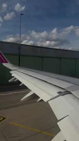 The video shows a view from an airplane window, looking out at the wing and the airport tarmac. The Wizz Air logo is visible on the tail of the wing. In the background, there is a green hangar-like building, a control tower, and another airplane with the Ryanair logo visible. The sky is blue with some white clouds. The sun appears to be shining. There are no people visible in the frame. The video appears to be taken while the plane is stationary at the gate or taxiing slowly.