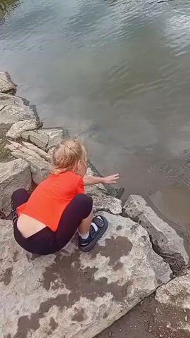 A young child with blonde hair, wearing an orange t-shirt and black leggings, squats on rocks near the edge of a body of water. The child is reaching out to touch the water. The water is murky and appears to have ripples. There are large rocks along the edge of the water. In the background, the water extends to the horizon.