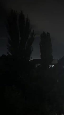 The video shows a dark outdoor scene at night. Two tall, slender trees are silhouetted against a dimly lit sky with scattered clouds. In the background, faint lights suggest the presence of buildings or houses. The overall impression is one of quiet darkness, possibly a rural or suburban setting. There are no people or overt actions depicted.