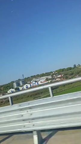 The video shows a view from a vehicle traveling on a road. A metal guardrail is prominently in the foreground. Beyond the guardrail, there is a landscape with green fields, some trees, and buildings. In the distance, several large industrial silos are visible, along with some smaller white buildings and what appear to be houses. The sky is a clear, bright blue. The video appears to be shot during daylight hours, likely morning or afternoon given the bright sunlight. There are no people visible in the video. The overall scene suggests a rural or semi-rural industrial area.