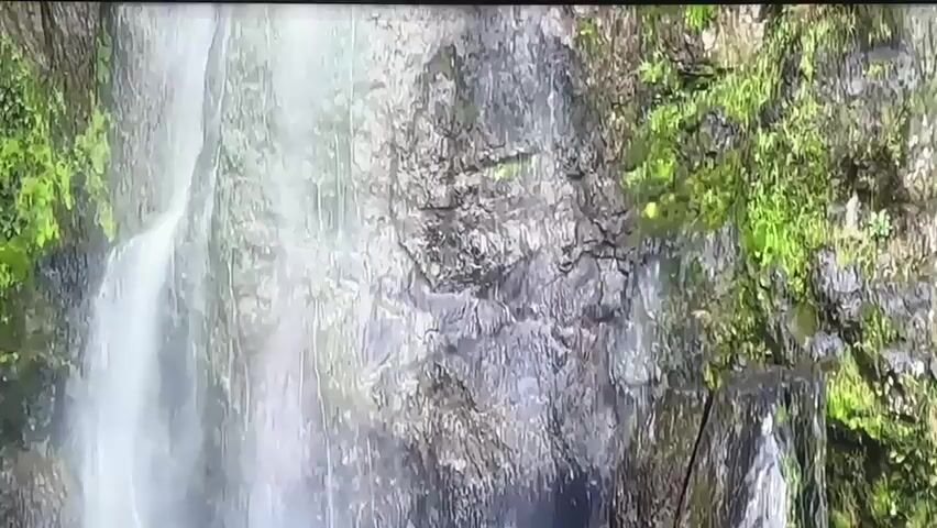 This is a close-up video of a waterfall cascading down a rocky, moss-covered cliff. The water is white and frothy as it descends, creating a blurred effect. The rocks are a mix of grey and brown, with patches of vibrant green moss clinging to them, especially on the right side of the frame. The scene appears to be in a natural, outdoor setting, likely a forest or mountainous area. There is no visible text in the media. The time of day and weather are not explicitly indicated, but the lighting suggests it is daytime with some natural light. The overall mood is serene and natural, emphasizing the beauty of the waterfall and its surroundings.