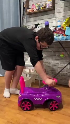 A fair-skinned person wearing glasses, a black long-sleeved shirt, and black shorts bends over and grabs the steering wheel of a purple toy car with pink accents. They appear to be attempting to move the car. The person is standing on a wooden floor in a room with a brick-patterned wall. Behind them is a white high chair, and a table with various items on top. The car has a license plate that reads "A911AA".