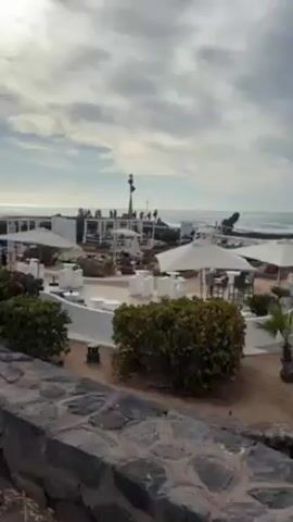 The video shows a beach club on a cloudy day. In the foreground is a stone wall with green bushes. Behind the wall is a mostly white-colored beach club setup, with several beach umbrellas, lounge chairs, and tables. In the background, people are walking or standing around on a raised deck area near the ocean. The ocean waves are visible, and the sky is mostly covered by clouds. Loud music plays in the background.
