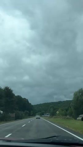 The video shows a view from inside a car driving on a paved road. The sky is overcast with gray clouds. On both sides of the road, there are trees and hills covered in foliage. A white car is visible ahead on the road, and there are road markings. There are also some buildings and signs visible in the distance. The overall impression is of a drive through a natural landscape under cloudy weather.