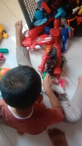 A young child is sitting cross-legged on a tiled floor surrounded by toys. The child has dark hair and is wearing a reddish-brown shirt and light-colored pants. He is facing a pile of toys, including a large red robot toy, a variety of smaller plastic toys, and some building blocks.  A black plastic basket full of toys sits to his left. The child is picking up and manipulating the toys, seeming to engage in imaginative play. The camera is positioned directly above the child, providing a bird's-eye view of the scene.