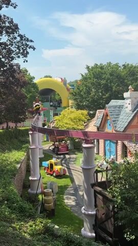 The video shows a theme park attraction. A small train or vehicle is on a track, surrounded by cartoon-like houses and trees. There are also what appear to be large inflatable structures or rides in the background. The weather is sunny with a blue sky and some clouds. The focus is on the colorful and whimsical environment of the park.