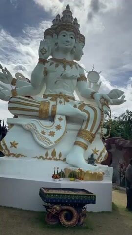 A video pans across an outdoor area featuring large statues. In the foreground, a statue of a multi-headed deity is seated, holding a mala. To its right, a statue of an elephant stands with its trunk lowered. Further back, a statue of a green-skinned, large figure with a sword is visible, with another elephant statue beside it. The scene then shifts to show a statue of a bird-like creature on a pedestal, with a large tree to its left. The video continues to pan, revealing a pathway with several people walking. A large elephant statue is prominent on the left, with a woman taking a photo of it. More people are seen walking along the path, with various statues and structures in the background. The environment appears to be a park or temple complex with lush greenery and cloudy skies.