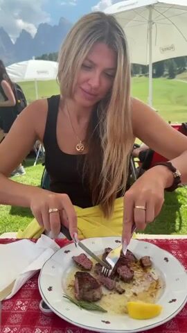 A woman with long blonde hair is eating a steak outdoors at a table covered with a red and white checkered tablecloth. She is wearing a black tank top and a yellow garment around her waist. She is using a knife and fork to cut the steak, which is on a white plate with a decorative rim. A sprig of rosemary and a lemon wedge are also on the plate. In the background, there are green hills, trees, and white umbrellas, suggesting a mountainous or scenic location. The woman is smiling and looking down at her food.
