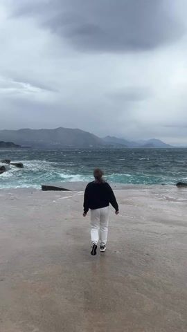 The video shows a woman walking toward the sea on a concrete pier on a stormy day. The woman is wearing a dark top, white pants, and athletic shoes.  The pier is wet. The sea is choppy with white-capped waves crashing against a rocky outcrop to the left of the woman. Mountains are visible in the background beneath a cloudy sky. The woman is seen from behind. She does not interact with anything during the video.