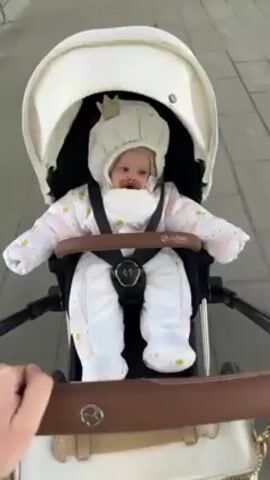 A baby wearing a white snowsuit with pink and gold details, a knit hat with a gold crown on top, and a pacifier is being pushed in a Cybex stroller with a white and brown frame on a paved surface. The baby's eyes are open and they are looking to the left. The stroller is being pushed by a person whose arm is visible in the foreground, holding the brown handle.