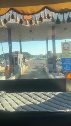 The video shows a view from inside a vehicle looking forward through a windshield. The vehicle is approaching a toll booth plaza. The camera view captures a straight, paved road with multiple lanes. On either side of the road are toll booths, each with a white booth structure and a red traffic light on a pole. In the distance, the road stretches towards a line of hills under a clear blue sky. As the vehicle moves forward, one of the traffic lights at the toll booth turns from red to green, indicating the path is clear. Cars are visible in other lanes, some waiting and some moving. The foreground of the video is dominated by the interior of the vehicle, including a quilted upholstery on a surface below the windshield and decorative tassels hanging from the top of the windshield.