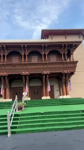 The video pans across the facade of a building with intricate wooden architectural details. The building features a wooden balcony with carved railings and columns. Below the balcony, there are several large wooden doors. American flags are displayed on flagpoles on either side of the entrance. Green carpeting covers a wide set of stairs leading up to the doors, with metal handrails on either side. Potted plants are placed along the sides of the stairs. The sky above is cloudy.