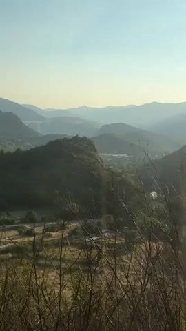 A drone shot pans across a valley filled with rolling green hills and scattered trees. A village is visible nestled in the valley, with a bridge spanning a river in the distance. The sky is a clear blue, with a few wispy clouds. The sun is setting, casting a warm glow over the landscape.