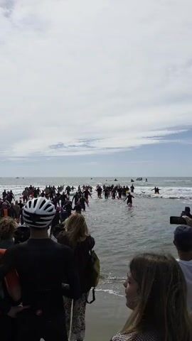 A medium shot captures a crowded beach scene during a race, with many participants in black wetsuits and pink caps wading through the waves. They are holding pink balloons and orange floats. The camera is positioned slightly behind the crowd on the shore, with a cyclist wearing a helmet partially visible in the foreground. Several spectators are also visible on the shoreline, one holding a smartphone to record the event. The sea is choppy, and the sky is overcast. In the background, there are jet skis, a boat, and two people on surfboards on the water.