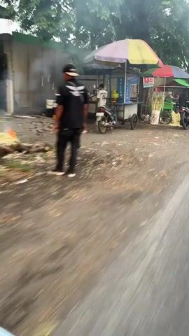 The video shows a street scene with several small food stalls. In the foreground, a road is visible with a blurred perspective, suggesting movement. To the left, there is a stall with a colorful umbrella, a motorcycle parked in front, and a person working at the stall. Sugarcane stalks and a sugarcane juicer are also visible at this stall. Further down the street, another stall with a green cart and an umbrella is seen, with a person attending to it. Behind this, a larger sugarcane juicing machine is visible. A pickup truck with a blue tarp is parked further down the road. The environment appears to be an outdoor market or roadside with trees and a wall in the background. The lighting suggests it is daytime, possibly late afternoon or early morning.