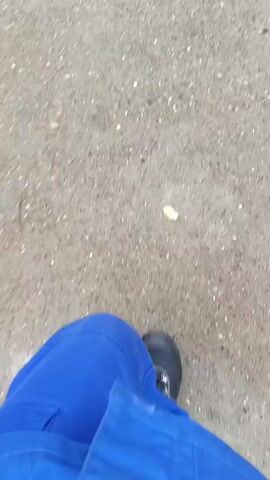 A first-person perspective video shows a person's legs and feet walking on a pavement. The person is wearing black boots and bright blue trousers with reflective silver stripes on the legs. The pavement appears to be concrete with small gravel mixed in, and there are some scattered green leaves and small pebbles. The camera is pointed downwards, focusing on the walking motion. The overall environment suggests an outdoor setting, possibly a sidewalk or path.