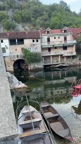 The video pans from a view of two small boats moored at the edge of a body of water, with a building complex situated on a hillside overlooking the water. The building is made of stone and has multiple stories with balconies and red tiled roofs. The water is calm and reflects the buildings and surrounding trees. Further down the waterway, a larger, red boat with a white canopy is also moored. The banks of the waterway are lined with trees and vegetation, and the overall impression is one of a tranquil, natural setting with a touch of human habitation. The video concludes with a wider view of the waterway extending into the distance, surrounded by lush green hills.