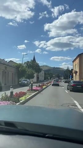 The video shows a view from inside a vehicle driving down a street. The street has a row of cars on it, and there are buildings and trees on either side. There is a prominent church spire in the distance, and hills can be seen beyond that. The sky is blue with white clouds. The video appears to be a recording of a car's journey through a town.