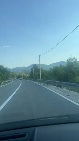 The video shows a car driving on a curved, paved road lined with green trees and vegetation. Mountains are visible in the distance under a clear, blue sky. The road has a white centerline and a metal guardrail on the right side. A few cars are visible ahead on the road. Power lines stretch across the sky on the right. The camera is positioned from the driver's perspective inside the car, capturing the dashboard's top edge.