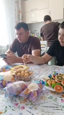 A man in a brown t-shirt is sitting at a table covered with a floral tablecloth, holding a smartphone in his hands and looking down at it. Beside him, another man, partially visible, is also seated at the table, reaching towards the camera. In front of them, on the table, are several packets of cookies and a plastic container filled with small, shell-shaped cookies. In the background, a woman with her hair tied back is visible in a kitchen area. The scene appears to be indoors, possibly a dining room, with a window providing natural light.