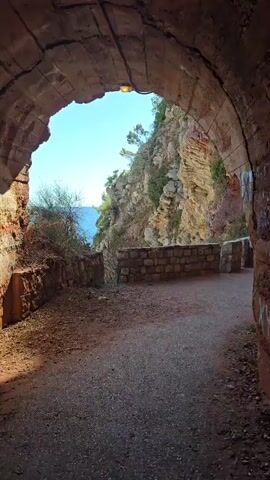 The video begins inside a dark tunnel with a rough, arched ceiling. A single light fixture is visible on the ceiling. The tunnel opens to reveal a bright, sunlit scene of a rocky cliff face overlooking a body of blue water. A dirt path leads from the tunnel towards the opening. A low stone wall follows the edge of the path. To the right of the path, inside the tunnel, a woman in a white dress stands looking out towards the water. Bushes and trees grow on the cliff face.