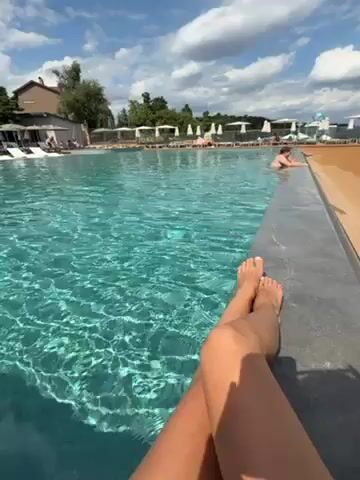 A low-angle shot shows a woman’s bare legs, crossed at the ankles, as she sits on the edge of a large, turquoise-colored swimming pool. The pool’s edge is made of gray tile. The pool extends into the distance, with several people visible in the water and along the sides. White lounge chairs with umbrellas are lined up along the edge of the pool. Beyond the pool, there are trees and a low, light-colored building. The sky is blue with scattered white clouds. The sun is shining, casting a bright light on the scene. The water in the pool is clear and reflects the sky. The legs are in focus and appear tanned.