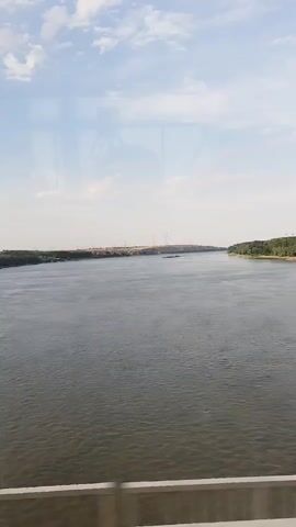 A wide, calm river flows under a bright, cloudy sky. The riverbanks are lined with trees on the right and a more barren, dusty landscape on the left, dotted with what appear to be power pylons. A small barge or boat is visible in the middle distance on the left side of the river, and a green buoy marks a channel further left. A white railing is visible at the bottom of the frame, suggesting the viewpoint is from a bridge or elevated structure. The overall scene depicts a serene, expansive natural environment.