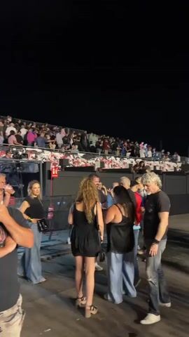 A group of people stand near a stage at night, with a large audience seated in bleachers behind them. The people in the foreground are casually dressed, with some wearing band t-shirts. One man in the front left is wearing glasses and a black t-shirt with the AC/DC logo. He has his arms crossed and is looking towards the right. To his right, a woman in a black top and denim jeans is looking at the man. Further right, a couple is talking, with the woman in a black dress and the man in a black t-shirt and jeans. Behind them, a man with grey hair is looking to the left. The audience in the bleachers is a diverse group of men and women of various ages, many of whom are looking towards the stage. The environment appears to be an outdoor venue, possibly a concert or festival, with temporary stage equipment and lighting visible. A Spanish flag is visible in the background behind the audience. The lighting is dim, with stage lights illuminating the foreground.