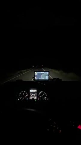 The video shows the interior of a vehicle at night, likely a truck, as seen from the driver's perspective. The dashboard is illuminated with glowing gauges and a GPS navigation screen displaying a map. The headlights of the vehicle cut through the darkness, illuminating a highway with faint lights visible in the distance. The scene conveys a sense of solitary travel during nighttime.