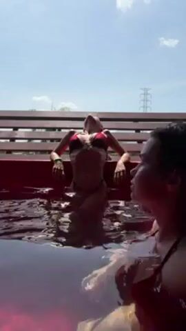 Two young women are in a red plastic tub, presumably a hot tub or small pool, with a dark liquid that appears to be water. The woman in the foreground has long dark hair that is wet, and she is wearing what looks like a red swimsuit or top. The other woman, further away, is also in the tub and seems to be wearing a dark bikini top. They are both looking out towards a wooden fence or railing. The sky is visible in the background, and there is a power line tower in the distance. The light indicates it is daytime. They are both relaxing in the tub.