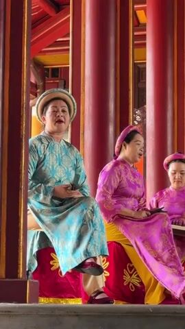 Three women in traditional Vietnamese dress are performing music. The woman on the left is wearing a blue and teal outfit and is singing. The two women to her right are wearing pink outfits and are playing musical instruments. The woman in the middle is playing a stringed instrument, possibly a dan tranh. The woman on the far right is playing a lute-like instrument. They are seated in front of a large, colorful mural depicting a bird, possibly a phoenix. The scene appears to be indoors, with large red columns and ornate architecture visible. The lighting is bright, suggesting daylight or stage lighting. The overall atmosphere is one of cultural performance and artistic expression.