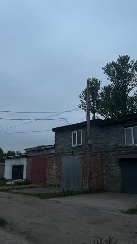 The video presents an exterior shot of a one-story building with multiple garage doors and a few windows, under an overcast sky.  The building appears to be constructed from cinder blocks, with portions featuring red brick and white brick. Utility poles and wires are visible in the foreground. Trees are situated behind the building, partially obscuring the sky. A black car is parked in front of one of the garage doors. The ground appears to be a mix of dirt and concrete.